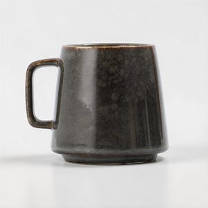 Puede incluir: Una taza de cerámica gris oscuro con una forma única, ligeramente acampanada. La taza tiene un asa robusta y una superficie texturizada, lo que le da un aspecto rústico. El interior y el borde son de un tono gris más oscuro.