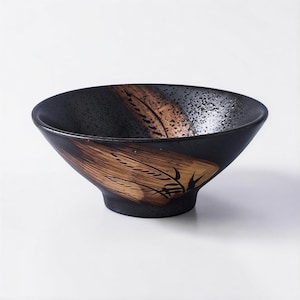 May include: A black ceramic bowl with a wide, angled design. The bowl features a brown and gold leaf-like pattern on the exterior. The interior has a speckled texture. The bowl is suitable for serving soups, salads, or other dishes.