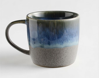 May include: A ceramic mug with a rounded body and a handle. The mug features a gradient of colours, transitioning from a dark green rim to blue and ending with a grey base. The mug is designed for hot beverages.
