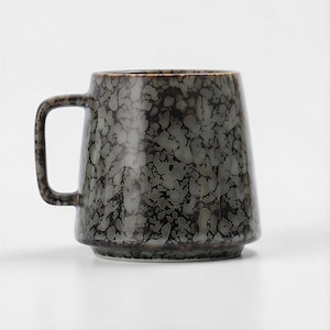 Puede incluir: Una taza de cerámica con un esmalte moteado gris oscuro y marrón. La taza tiene un cuerpo ancho, ligeramente cónico, un asa robusta y una base elevada. El interior es de color gris claro.