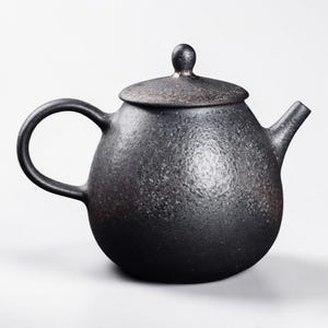 Op de afbeelding: Zwarte keramische theepot met een afgeronde body, een gebogen handvat en een tuit. Het deksel heeft een kleine knop bovenop. De theepot heeft een getextureerd oppervlak en een matte afwerking.