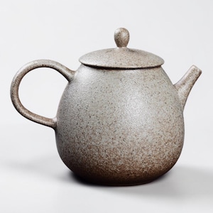 Puede incluir: Una tetera de gres moteado con cuerpo redondeado, asa curva y boquilla. La tapa tiene un pequeño pomo. La tetera es de color neutro con motas marrones, adecuada para preparar té.