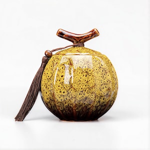 Op de afbeelding: Een decoratieve keramische pot met een deksel en kwast. De pot is rond met een geel en bruin gespikkeld patroon. Het deksel heeft een bruin handvat en een bruine kwast hangt aan de zijkant. De pot wordt waarschijnlijk gebruikt voor opslag of decoratie.