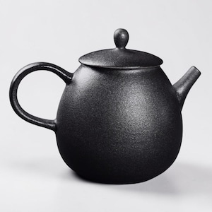 Op de afbeelding: Een zwarte keramische theepot met een getextureerd oppervlak. De theepot heeft een afgeronde body, een gebogen handvat, een tuit en een deksel met een kleine knop. De theepot is ontworpen voor het zetten van thee.