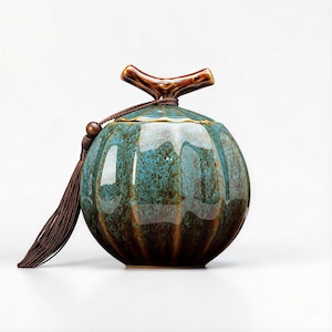 May include: A round, ceramic jar with a teal and brown speckled glaze. The jar has a lid with a brown branch-shaped handle and a brown tassel. The jar is a decorative item.