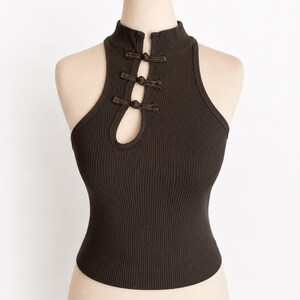 May include: A dark brown sleeveless top featuring a ribbed texture, a high mock neckline, and a keyhole cutout at the chest. The garment is secured with three decorative knotted frog closures aligned vertically along the centre front opening.