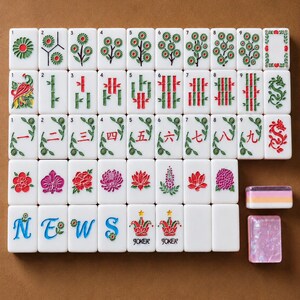 May include: A set of white rectangular Mahjong tiles featuring colorful engraved designs including flowers, bamboo stalks, Chinese numerals, the word NEWS, and Joker tiles with stars. Two additional tiles are placed separately to the right.