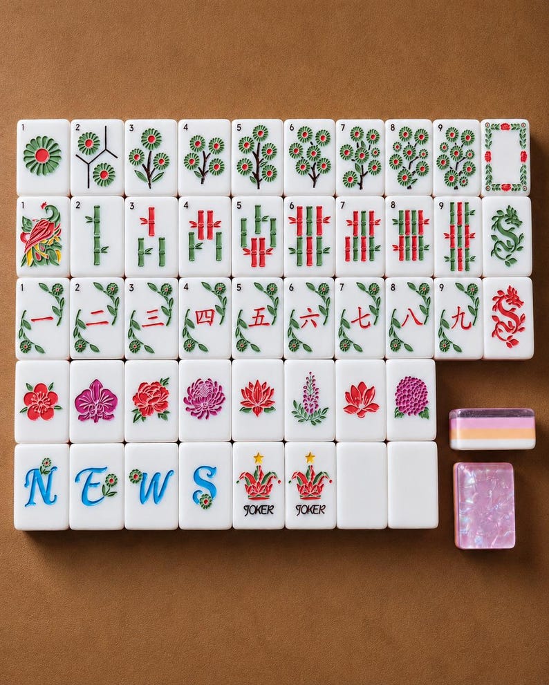 May include: A set of white rectangular Mahjong tiles featuring colorful engraved designs including flowers, bamboo stalks, Chinese numerals, the word NEWS, and Joker tiles with stars. Two additional tiles are placed separately to the right.