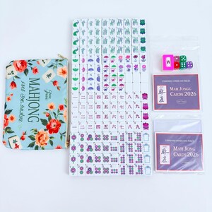 May include: A Mahjong set featuring a light blue floral storage pouch, a complete collection of white tiles with colorful symbols, numbers, and characters, four small dice, and two packs of 2026 standard hands and rules playing cards.