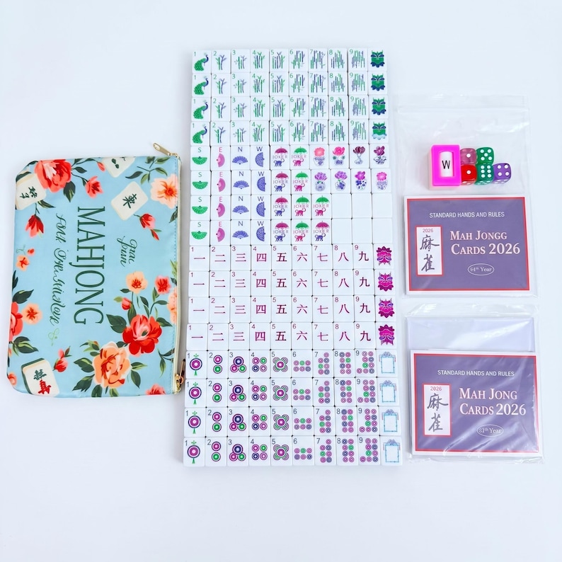 May include: A Mahjong set featuring a light blue floral storage pouch, a complete collection of white tiles with colorful symbols, numbers, and characters, four small dice, and two packs of 2026 standard hands and rules playing cards.