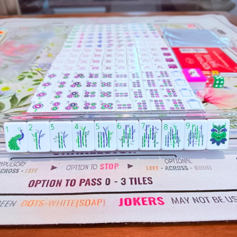 May include: A Mahjong game set featuring white tiles with green and purple ink designs, including numbered bamboo stalks, a peacock, and a dragon. The set includes a play mat with printed text instructions and rules for passing tiles during gameplay.