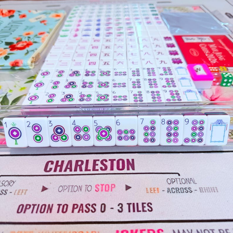 May include: A Mahjong set featuring white tiles with purple and green circular patterns, numbers one through nine, and a blank tile. The set includes a floral pouch, a 2026 card, and a game mat with the text CHARLESTON, OPTION TO STOP, and OPTION TO PASS 0-3 TILES.