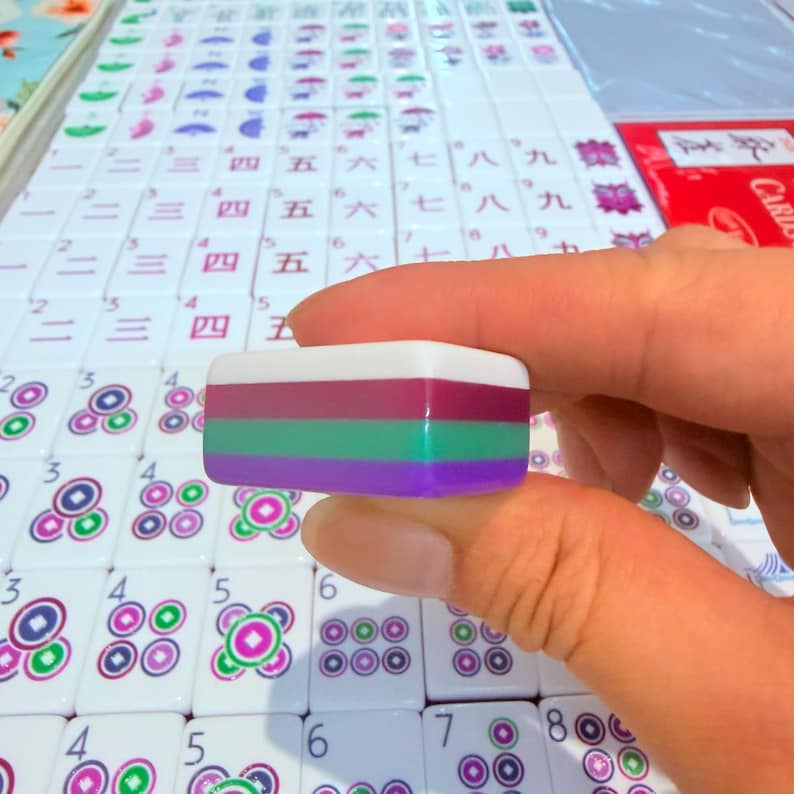 May include: A rectangular mahjong tile featuring a tri-color design with a white top layer, a magenta middle layer, a green center layer, and a purple bottom layer, held above a set of white mahjong tiles with various printed symbols and numbers.