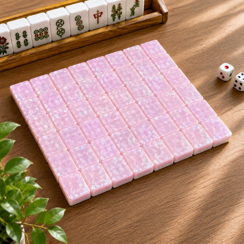 May include: A set of rectangular mahjong tiles with a shimmering, pearlescent pink finish arranged in a square grid. The tiles feature a smooth, reflective surface texture. Two white six-sided dice with black pips are positioned nearby.