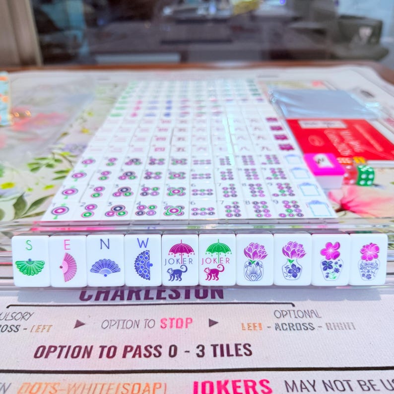 May include: A Mahjong set featuring white tiles with colorful printed designs including fans, monkeys under umbrellas labeled JOKER, and floral vases. The set is arranged on a game mat with text including CHARLESTON and OPTION TO PASS 0-3 TILES.