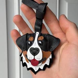 May include: A handmade leather dog tag in the shape of a Bernese Mountain Dog's head. The tag features black, brown, white, and red leather pieces stitched together. It has a black leather loop for attaching to a bag or keychain.