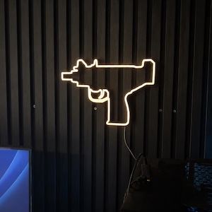 May include: A warm white neon light sign shaped like a submachine gun silhouette. The glowing outline features a barrel, trigger guard, and stock, mounted against a dark vertical slat wall. A thin power cord hangs from the bottom of the fixture.