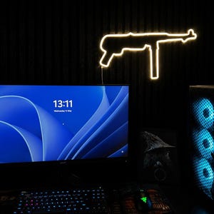 Puede incluir: Un contorno amarillo neón de una ametralladora está montado en una pared oscura. Un monitor de ordenador muestra un fondo abstracto azul y blanco. Un teclado, un ratón y una torre de ordenador son visibles en el escritorio de abajo.