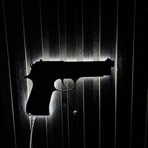 Puede incluir: Una silueta negra de una pistola, iluminada desde atrás, montada sobre una superficie oscura con líneas verticales. El contorno del arma está bien definido, con un gatillo y una empuñadura visibles. Un fino cable blanco sale de la parte inferior de la pistola.