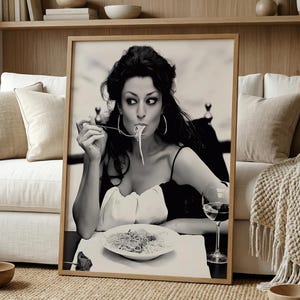 May include: Black and white framed print of a woman eating spaghetti. The woman wears a black and white dress and large hoop earrings. A glass of red wine and a plate of pasta are on the table. The print is displayed in a living room.