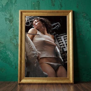 May include: A framed print featuring a person in a white tank top and underwear. The gold-colored frame contrasts with the green wall and brown wooden floor. The image has a vintage aesthetic.