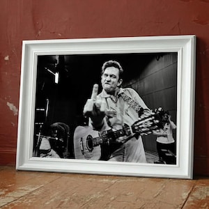 May include: A framed black and white photograph of a musician playing a guitar and giving the middle finger. The white frame rests on a wooden floor against a red wall. The image captures a moment of defiance.