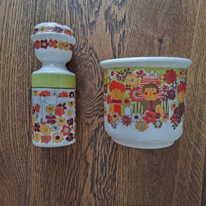May include: A white ceramic shaker and a matching bowl featuring a colorful floral pattern in shades of red, orange, and yellow. The shaker has a green band and a bow graphic, while the bowl displays stylized figures amidst the floral design.