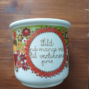 May include: White ceramic planter with a colorful floral design in orange, red, yellow, and brown. The planter has a green rim and a central text in a decorative frame. The text reads "Gild på mange vis til vertinnens pris."