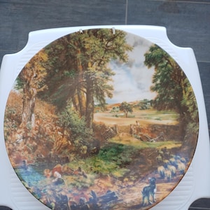 May include: Decorative plate featuring a pastoral landscape scene with trees, a stream, and sheep. The plate has a white rim and is held by metal clips. The artwork depicts a sunny day with figures and animals.