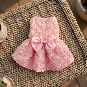 Peut inclure: Une robe rose clair pour chien avec un corsage textur&eacute; en forme de roses et un grand n&oelig;ud en satin. La robe a une jupe &eacute;vas&eacute;e et est con&ccedil;ue pour un petit chien. Le style g&eacute;n&eacute;ral est &eacute;l&eacute;gant et f&eacute;minin.