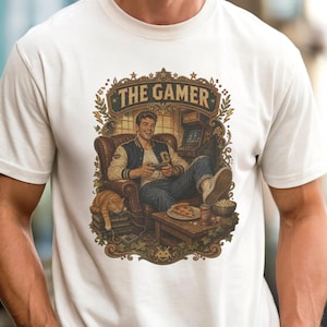 Op de afbeelding: Een ivoorkleurig t-shirt met een grafische illustratie van een persoon die in een leren fauteuil zit en een videogame speelt. Het ontwerp bevat de tekst THE GAMER, een kat, een stuk pizza en een kom met snacks, omlijst door decoratief blad.