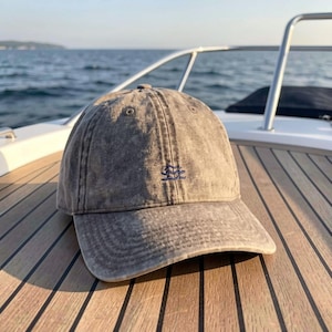 May include: A grey acid-wash baseball cap featuring a small, embroidered dark blue wave icon on the front centre. The cap has a structured crown with ventilation eyelets and a curved brim, showcasing a weathered, textured fabric appearance.