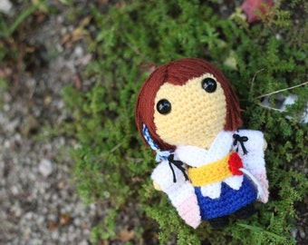 May include: Crocheted doll with brown hair, yellow skin, and large black eyes. The doll is wearing a white, pink, and blue outfit with a yellow sash.