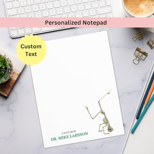 May include: White personalized notepad with a skeleton illustration. The notepad has the text "Personalized Notepad" at the top and "A note from Dr. Mike Larsson" at the bottom. A yellow scalloped shape with the words "Custom Text" is in the upper left corner.