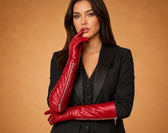 May include: A woman wearing a black suit and long red leather gloves with silver zippers. The gloves extend past the elbows. The background is a solid, warm brown color.