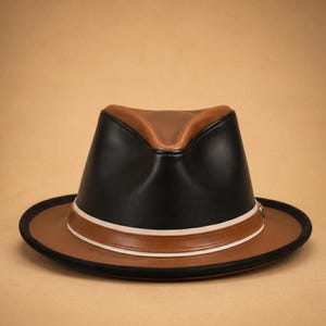 Puede incluir: Sombrero fedora de dos tonos con ala y corona marrones, y parte superior negra. El sombrero presenta una banda blanca y marrón alrededor de la base de la corona. Un accesorio elegante.