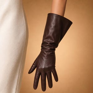 以下が含まれることがあります： 長いダークブラウンの革手袋。手袋は腕に沿って伸び、滑らかで光沢のある仕上がりです。指はよく定義されており、手袋は高品質の革で作られているようです。背景は暖かく、茶色です。