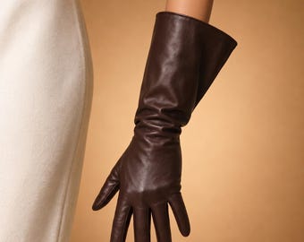 May include: A long, dark brown leather glove. The glove extends up the arm, with a smooth, polished finish. The fingers are well-defined, and the glove appears to be made of high-quality leather. The background is a solid, warm tan color.