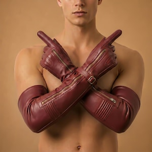 May include: Burgundy leather gloves with long sleeves, featuring gold zippers and a buckle closure. The gloves are designed with ribbed detailing and are shown with crossed arms.