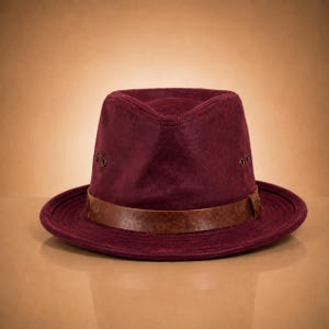 Puede incluir: Un sombrero fedora burdeos con una banda de cuero marrón. El sombrero tiene una corona estructurada y un ala ancha, ligeramente levantada. El sombrero está hecho de un material suave similar al ante.