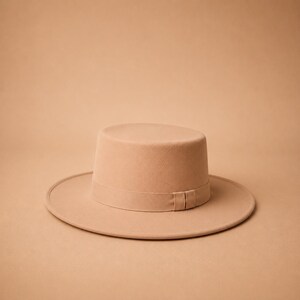 May include: A solid light tan hat with a flat crown and a wide, flat brim. The hat features a matching tonal ribbon band encircling the base of the crown, which includes a small, subtle knot detail on the side of the band for a clean finish.