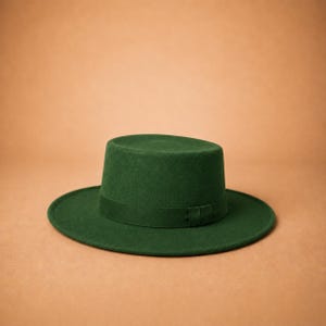 May include: A forest green flat-top hat with a wide, structured brim and a matching tonal ribbon band encircling the base of the crown. The hat features a smooth, felt-like texture and a clean, symmetrical silhouette with a flat top crown.