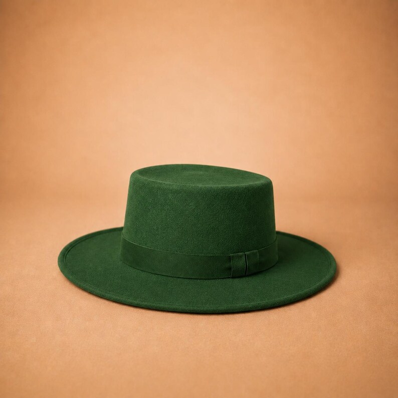 May include: A forest green flat-top hat with a wide, structured brim and a matching tonal ribbon band encircling the base of the crown. The hat features a smooth, felt-like texture and a clean, symmetrical silhouette with a flat top crown.
