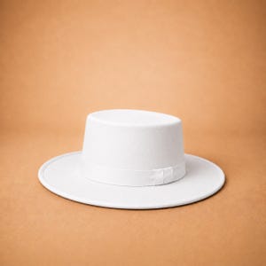 May include: A solid white hat featuring a flat crown and a wide, flat brim. The hat includes a matching white ribbon band encircling the base of the crown, providing a clean, uniform appearance with a structured shape and a smooth, matte finish.