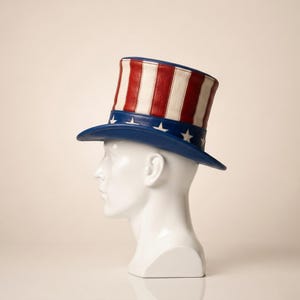 May include: A patriotic Uncle Sam top hat. The hat features a blue brim with white stars, a red and white striped crown, and a blue band. The hat is displayed on a white mannequin head.