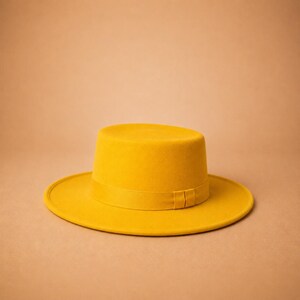 May include: A mustard yellow flat-brimmed hat with a structured cylindrical crown and a matching tonal ribbon band encircling the base. The hat features a wide, flat brim and a smooth, uniform felt-like texture throughout the entire surface.