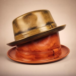 May include: A metallic gold fedora hat with a dark burnished finish rests atop a tan leather fedora hat. The tan hat features a matching leather band and a small embossed logo on the side. Both hats have a structured brim and crown.