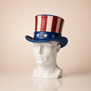May include: A patriotic Uncle Sam top hat. The hat features a blue brim with white stars, a red, white, and blue striped crown, and a blue band. The hat is displayed on a white mannequin head.