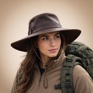 May include: A dark brown leather wide-brimmed hat featuring a structured crown, a matching leather hat band, and two brass-toned metal eyelets on the side. The hat includes a thin, adjustable leather chin strap with a sliding toggle for secure fit.