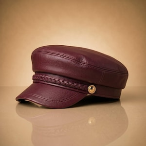 May include: A deep burgundy leather baker boy cap featuring a braided leather trim across the front. The hat includes a short, stiff brim and a polished gold-tone circular button accent on the side, providing a structured and classic appearance.
